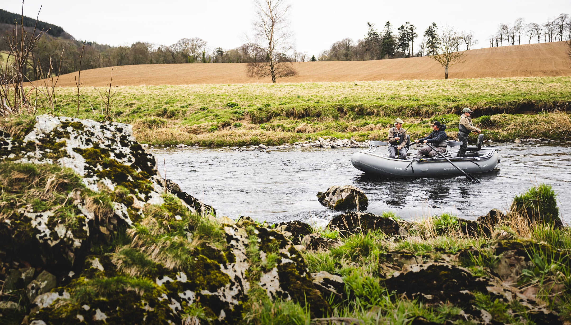 Three people on an inflatable boat in a river in scotland surrounded by grassy banks and trees.