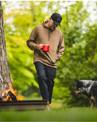 Man in a brown hoodie and black cap holding a red mug, walking with a dog near a campfire in a forest.