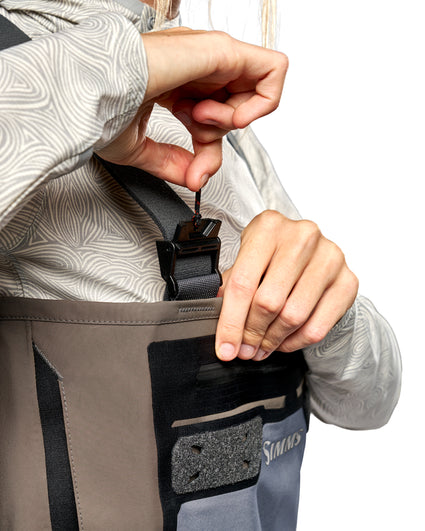 Women's Waders G3 Pro Guide Stockingfoot Close-up of a person wearing gray and black waders, adjusting the front buckle strap with both hands against a white background.