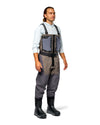 Men's G3 Pro Bootfoot Waders A man wearing a pair of gray and taupe waders with a black harness and chest pocket, a light blue long-sleeve shirt, and black rubber boots, standing against a white background.
