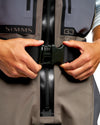 Men's G3 Pro Bootfoot Waders Close-up of a person wearing a Simms G3 waterproof jacket, fastening the black buckle closure over a black zipper. The jacket is gray and taupe with black details.