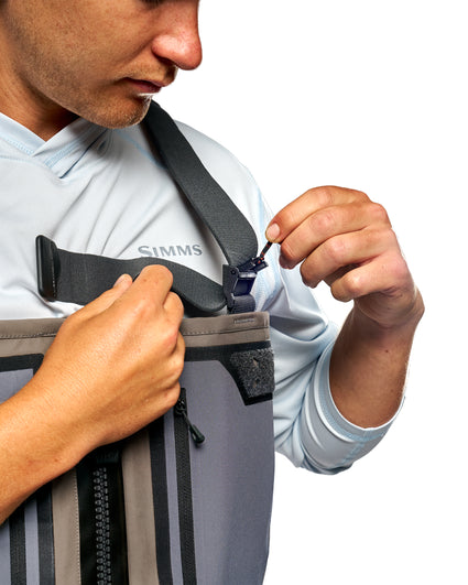 Men's G3 Pro Bootfoot Waders A close-up of a man wearing a gray and black Simms chest rig and hook-and-loop strap system, demonstrating how to buckle the strap. The Simms logo is visible on his light blue shirt.