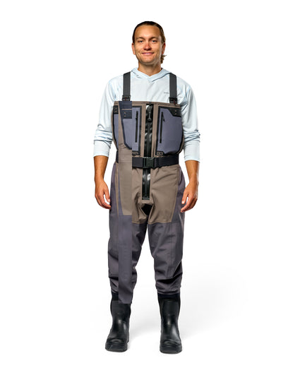 Men's G3 Pro Bootfoot Waders A man wearing a pair of gray and brown waterproof waders over a light blue hoodie and black rubber boots, standing against a solid white background.