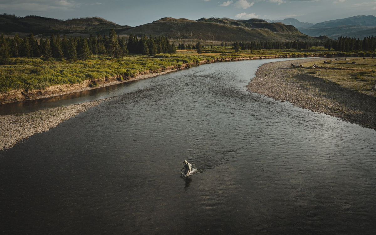 A wide aerial view of a river winding through a grassy landscape with mountains in the background, and a small person standing in the water near the center.