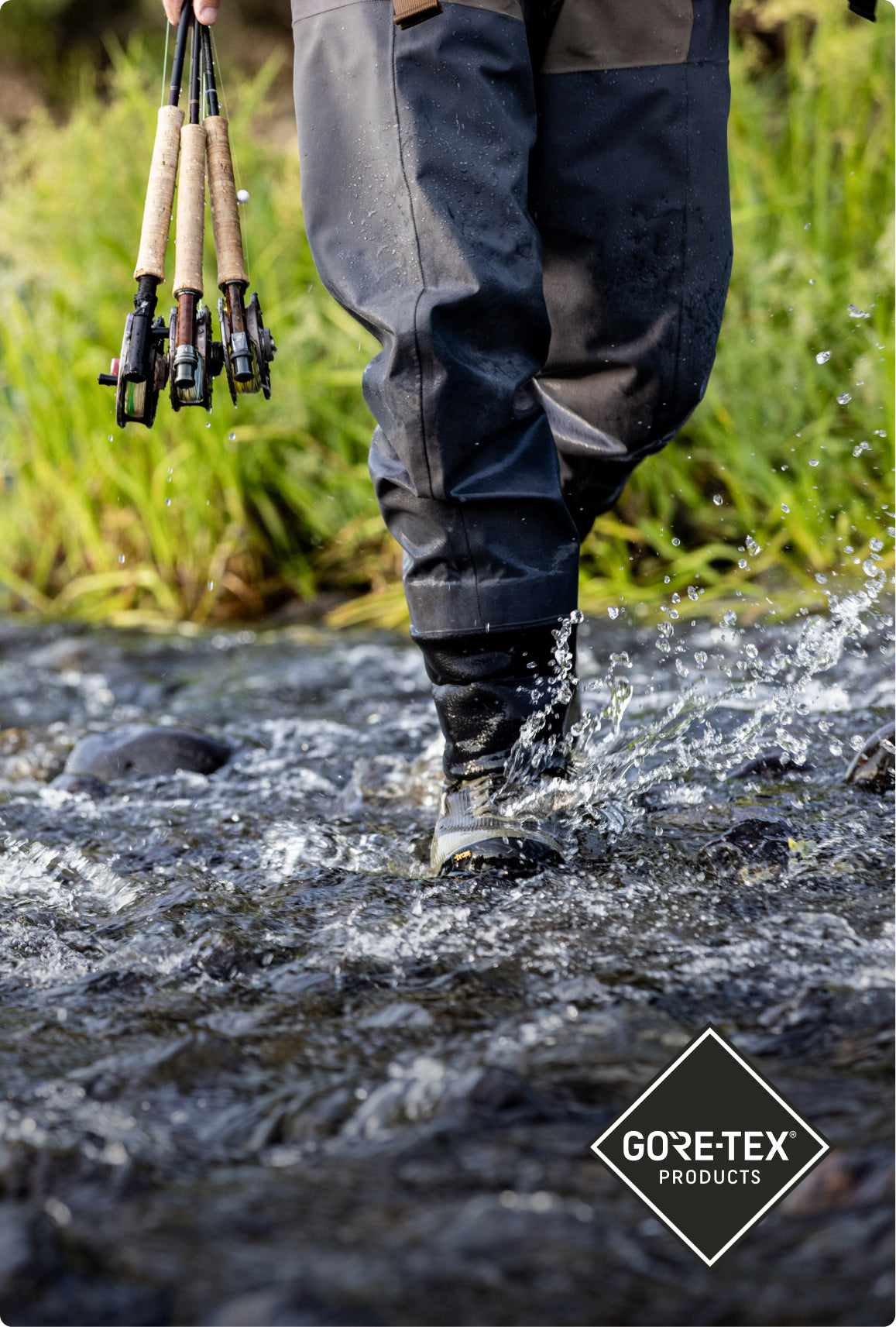 A person wading through a shallow stream wearing dark gray and black fly-fishing waders, holding three fishing rods. A white Gore-Tex Products logo is visible in the bottom right corner.