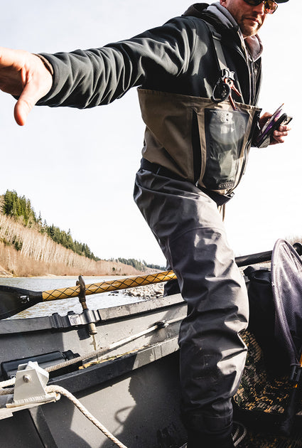 A man wearing dark gray and olive green fishing waders stands in a boat on water, holding a phone and pointing toward the left. The background shows a shoreline with trees and grass.