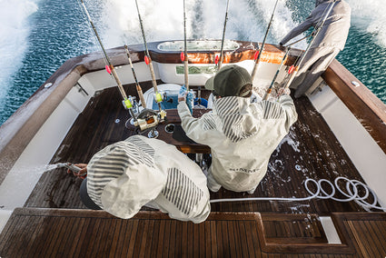Three people in white hooded Rip Line Pro rain suits work on the deck of a boat, with fishing rods, water, and blue ocean visible. The suits have a striped pattern and one person has 'CLIMB' printed on the back.