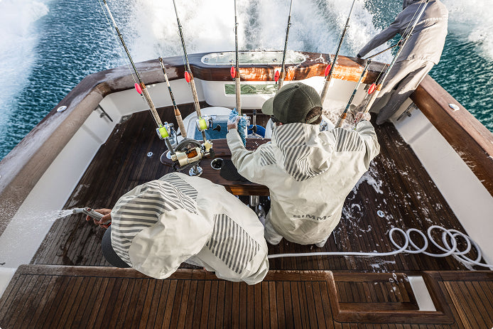 Three people in white hooded Rip Line Pro rain suits work on the deck of a boat, with fishing rods, water, and blue ocean visible. The suits have a striped pattern and one person has 'CLIMB' printed on the back.