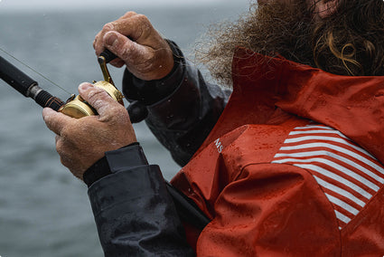 A close-up lifestyle shot of a bearded fisherman in an orange and white striped simms rip line pro waterproof jacket, holding a gold spincast fishing reel and rod against an overcast, misty water background.