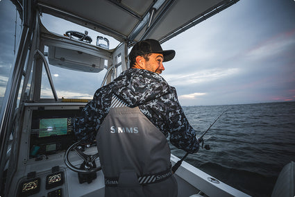 A man wearing a black cap and a gray simms rip line waterproof bib sits inside the cabin of a boat, facing the open water. He is wearing gray life jackets with the brand name 'SIMMS' printed on the back.