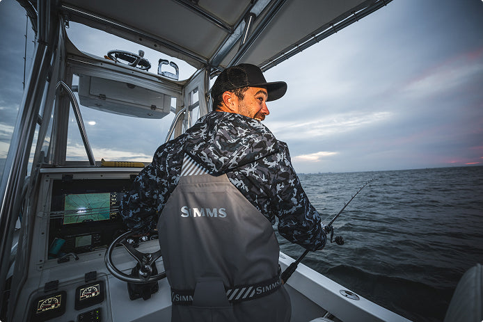 A man wearing a black cap and a gray simms rip line waterproof bib sits inside the cabin of a boat, facing the open water. He is wearing gray life jackets with the brand name 'SIMMS' printed on the back.