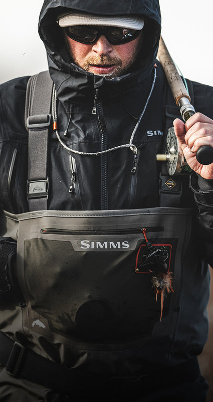A close-up of a person wearing a black Simms hooded jacket and dark chest rigging gear, holding a fishing rod. The Simms logo is visible on the chest piece and sleeve.