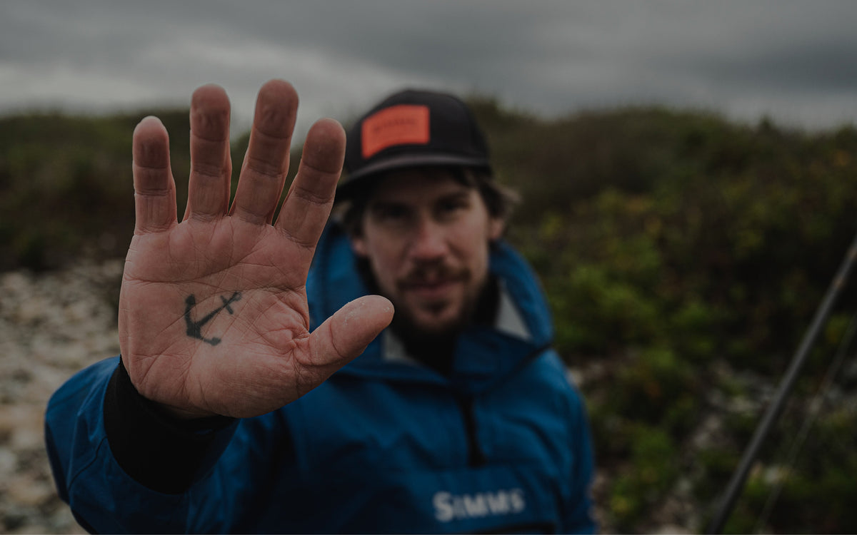 A man wearing a blue Simms jacket and a black cap with an orange label holds up his hand, showing a small anchor tattoo on the palm. The background is a blurred outdoor landscape with greenery and an overcast sky.