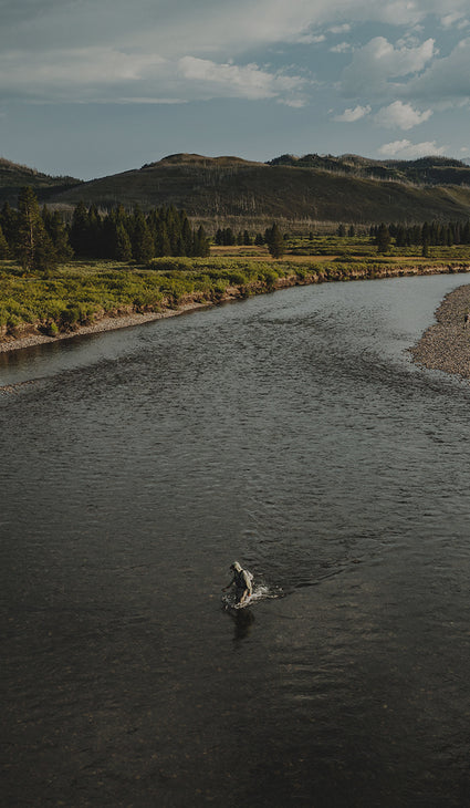 A wide outdoor landscape photo showing a river flowing through a grassy valley with hills and a cloudy sky. A small person is visible standing in the water near the lower center of the frame.