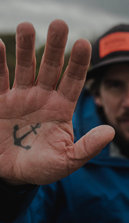 A close-up of a person's palm showing a small black anchor tattoo, with the person's face and a blue jacket blurred in the background.
