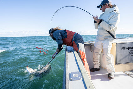 Two people on a boat in the open ocean, with one actively handling a fish using a net and the other standing nearby holding a fishing rod. The water is blue-green, the sky is clear, and part of a boat sign reading 'Newport' is visible on the side. They are wearing simms rip line waterproof fishing rain suits in gray and orange.