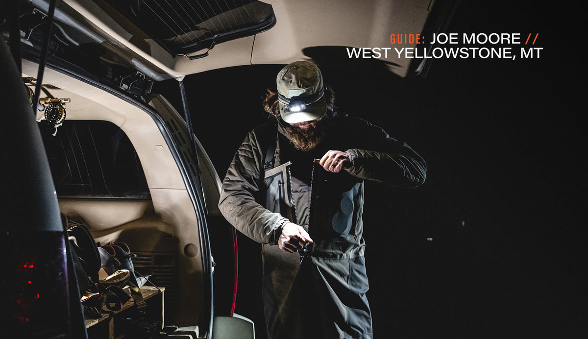 A bearded man wearing a camouflage cap and dark jacket stands in the open trunk of a vehicle at night, illuminated by a headlamp. Text in the top right reads 'GUIDE: JOE MOORE // WEST YELLOWSTONE, MT'.