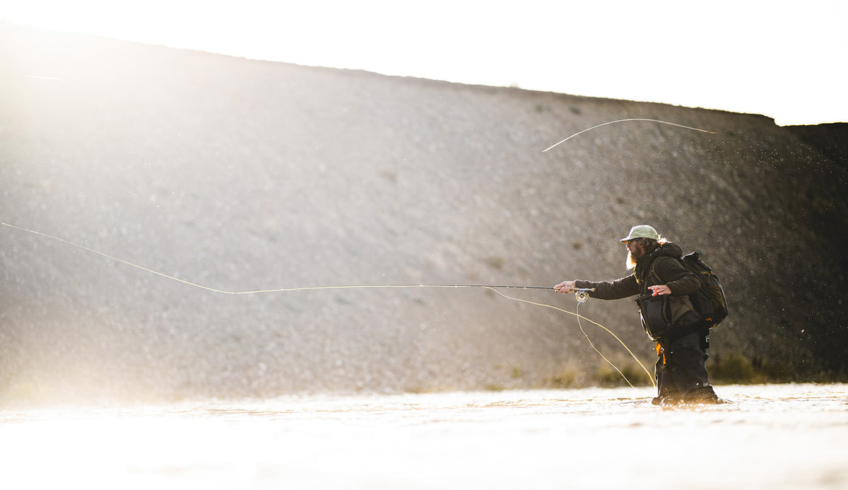 A person fly fishing in shallow water, casting a line with a fly rod, set against a large dark rock wall and bright, sunlit background.